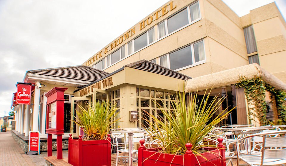 Beige hotel exterior with rooftop signage, red planters, spiky plants, and an outdoor café seating area.