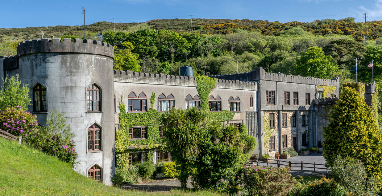 Gray crenellated castle-like building with ivy, gardens, flags, and wooded hills under a blue sky.